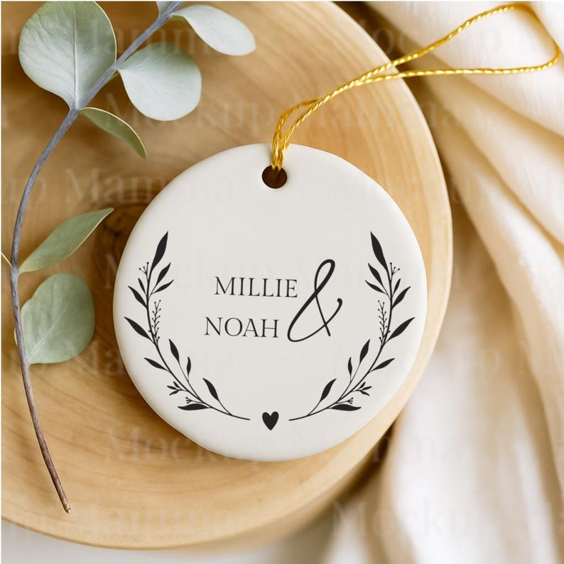 May include: A white ceramic ornament with the names "Millie & Noah" in black script, framed by a black floral wreath. A gold string is attached for hanging. The ornament is on a wooden surface.