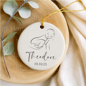 May include: A round, off-white ceramic ornament with a black line drawing of a sleeping baby. The name "Theodore" is written in cursive below the image, with the date "10.10.25" beneath it. A gold string is attached for hanging. The ornament rests on a wooden surface.