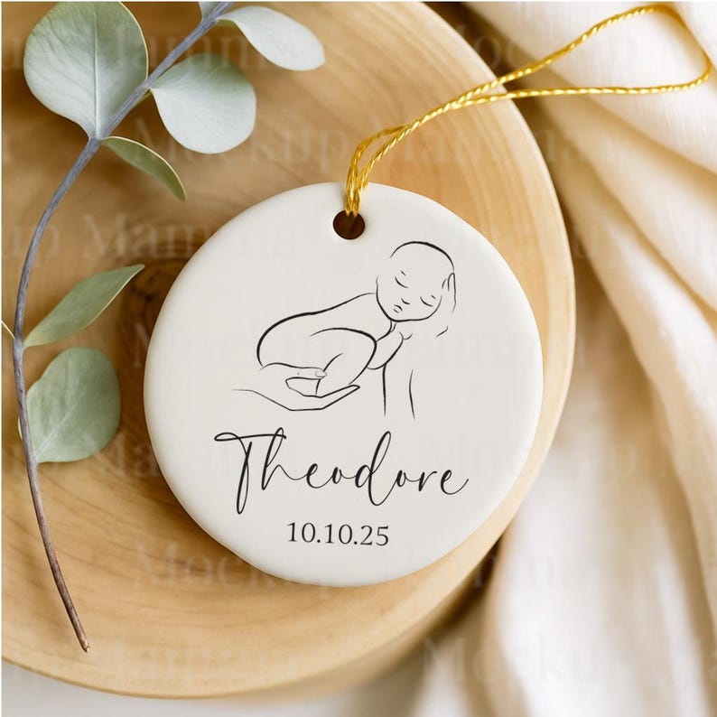 May include: A round, off-white ceramic ornament with a black line drawing of a sleeping baby. The name "Theodore" is written in cursive below the image, with the date "10.10.25" beneath it. A gold string is attached for hanging. The ornament rests on a wooden surface.
