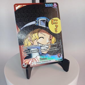 May include: A collectible trading card featuring Sabo from the Dressrosa/Revolutionary Army. The card has a holographic design with a portrait of Sabo, the text "Sabo" and "5000". The card is displayed on a black stand.