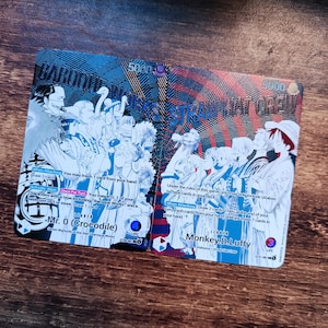 Może przedstawiać: Dwie karty kolekcjonerskie One Piece przedstawiające Mr. 0 (Crocodile) i Monkey D. Luffy. Karty zawierają ilustracje postaci, tekst gry, poziomy mocy 5000 oraz liczniki życia. Lewa karta pokazuje Baroque Works, a prawa Straw Hat Crew.