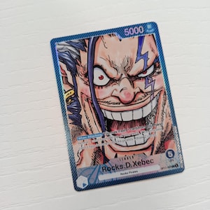 May include: A rectangular trading card featuring a colorful illustration of the character Rocks D. Xebec with a wide, grinning expression. The card displays the text Rocks D. Xebec, 5000 power, 5 life, and game instructions in blue and white.