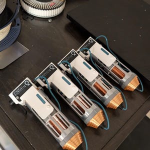 May include: Four white and gray devices with copper-colored ends and blue wiring are arranged on a dark wooden surface. Each device has a rectangular design with a series of parallel lines and a small blue rectangle. The devices appear to be electronic components.