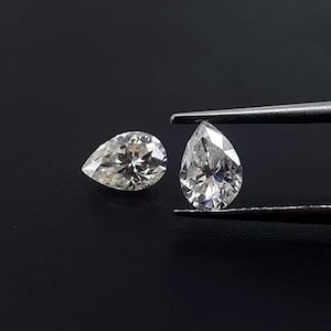 May include: Two pear-shaped diamonds on a black background. One diamond is held by tweezers, showcasing its facets. The other diamond is positioned to the left, also displaying its cut and clarity. The diamonds appear to be colorless.