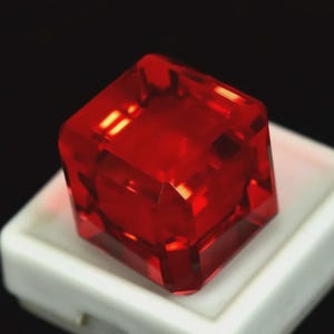 May include: A faceted, square-cut, red gemstone. The gem is displayed on a white surface against a black background. The gemstone's facets reflect light, creating a brilliant appearance. The gem is likely a ruby or a similar precious stone.