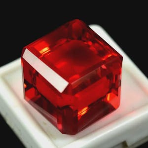 Pode incluir: Uma pedra preciosa vermelha vibrante, lapidada em forma quadrada e facetada. A gema está colocada sobre uma superfície branca com um fundo preto. As facetas da gema refletem a luz, criando uma exibição brilhante de cor e clareza. A gema tem a forma de um cubo.