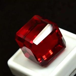 May include: A faceted, deep red gemstone with a square shape. The gem is displayed on a white surface against a black background. The cut of the stone creates multiple reflective surfaces.