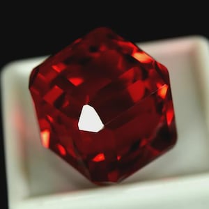 Peut inclure: Une pierre précieuse rouge foncé à facettes, de forme carrée. Les facettes de la gemme reflètent la lumière, créant un effet scintillant. La pierre est posée sur un fond blanc.
