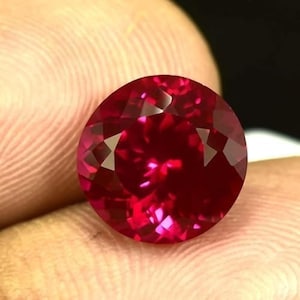 May include: A faceted, round-cut ruby gemstone. The gem is a deep, saturated red color with internal reflections. The ruby is held between fingers, showcasing its brilliance and clarity.