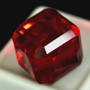 May include: A faceted, deep red gemstone with a square shape. The gem has multiple reflective surfaces, creating a brilliant display of light and color. The gemstone is set against a white surface.