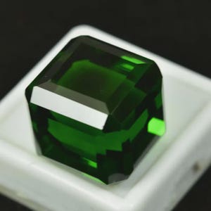 Puede incluir: Una gema verde esmeralda facetada de forma cuadrada. La gema está colocada en una bandeja blanca sobre un fondo oscuro. Las facetas reflejan la luz, creando una exhibición brillante de color y claridad. Ideal para la creación de joyas.