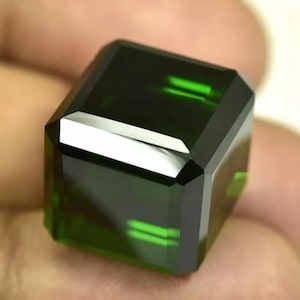 May include: A faceted, emerald-green gemstone with a rectangular shape. The gem has sharp, clean edges and a polished surface that reflects light. The deep green color is consistent throughout the stone, with subtle internal reflections.