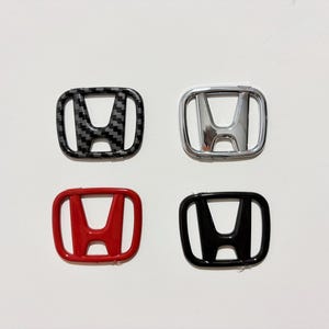 May include: Four Honda emblem badges in a square arrangement. The badges include carbon fiber, chrome, red, and black finishes. Each badge features the Honda "H" logo within a rectangular frame.