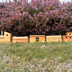 May include: A wooden toy train set with five cars, each with wheels. The train is made of light-colored wood and is sitting on green grass. The background features a bush with red and purple leaves.
