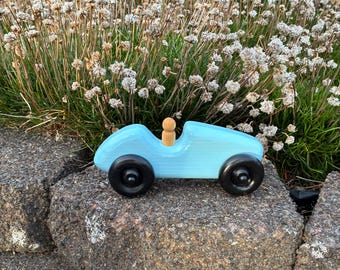 May include: A light blue wooden toy car with black wheels and a small wooden peg person in the driver's seat. The toy car is sitting on a large gray rock. The background features a cluster of small white flowers.