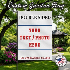 May include: A white, double-sided garden flag with the text "YOUR TEXT/PHOTO HERE" in red. The flag also features the text "DOUBLE SIDED" and "FLAG STANDS ARE NOT INCLUDED". The flag is displayed outdoors in a garden setting.