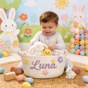 May include: A white rope basket with the name "Luna" embroidered in purple, decorated with fabric flowers. The basket contains a plush white bunny and a yellow chick. A small white bunny figurine and colorful Easter eggs are nearby.