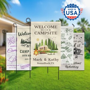 May include: A collection of personalized garden flags with camping themes. The flags feature illustrations of campsites, RVs, and text such as "Welcome to our Campsite" and names. The flags are displayed outdoors on metal stakes.