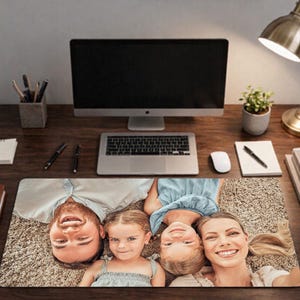 May include: A rectangular desk mat featuring a high-resolution color photograph of a family of four lying on a textured carpet. The image displays a man, a woman, and two young children smiling at the camera from an overhead perspective.