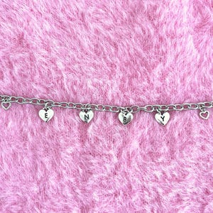 Könnte beinhalten: Eine silberfarbene Kette mit herzförmigen Anhängern. Die Anhänger buchstabieren "S E N B Y" mit einzelnen Buchstaben. Die Kette wird vor einem rosa, flauschigen Hintergrund präsentiert.