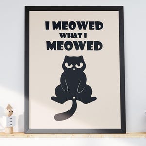 May include: A framed print with the text "I Meowed What I Meowed" and a black cat illustration. Above, four framed botanical prints hang on the wall. A shelf below holds decor, including books, candles, and a small plant.