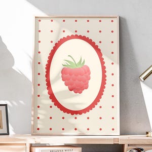 Puede incluir: Un estampado enmarcado con una ilustración de frambuesa dentro de un óvalo rojo sobre un fondo crema con lunares rojos. El estampado se exhibe en un escritorio de madera con una pequeña planta, libros y una lámpara de escritorio dorada. El tema general es la decoración del hogar.