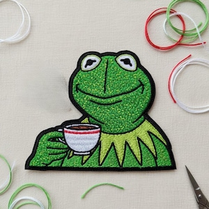Può includere: Toppa ricamata raffigurante una rana verde che tiene in mano una tazza di caffè. La rana sorride ed è bordata di nero. La tazza è bianca con una striscia rossa. Fili verdi, bianchi e rossi e piccole forbici sullo sfondo.