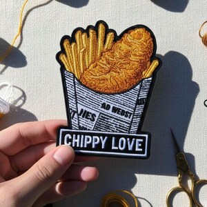 Pode incluir: Patch bordado com uma caixa de batatas fritas douradas e um bife de frango frito, com o texto "CHIPPY LOVE" num retângulo branco. O patch é preto, branco e amarelo. O fundo inclui bobinas de fio amarelo, branco e castanho.