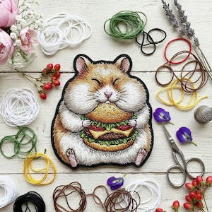 Op de afbeelding: Geborduurde patch met een hamster die een hamburger eet. De patch is bruin, wit en zwart met gedetailleerde steken. De afbeelding bevat bloemen, draad, een schaar en een vingerhoed, wat wijst op een knutselthema.