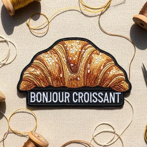 May include: Embroidered croissant patch with the text "BONJOUR CROISSANT". The patch is surrounded by spools of thread and gold scissors. The image has a craft theme.