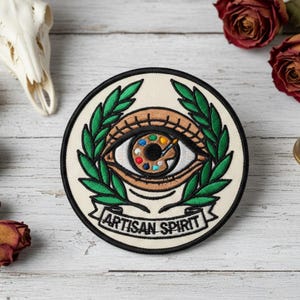 May include: Circular patch with an eye design, featuring a palette of colors within the iris and a paintbrush. The eye is framed by green laurel leaves, with the words "ARTISAN SPIRIT" on a banner below. The patch has a black border.