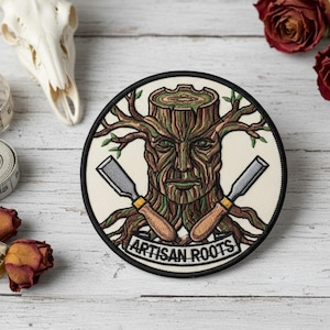 May include: Circular patch with a tree-like face design, featuring a tree trunk with a face, branches, and crossed chisels. The text "ARTISAN ROOTS" is below the face. The patch has a black border and is on a cream background.