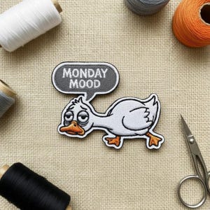 May include: Embroidered patch of a white duck with a weary expression, orange beak and feet. A grey speech bubble above the duck reads "MONDAY MOOD". The patch is surrounded by spools of thread and a pair of scissors.
