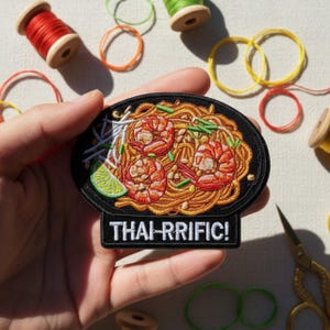 Puede incluir: Parche bordado que representa Pad Thai con gambas, fideos y lima. El parche es negro con la inscripción "THAI-RRIFIC!" en blanco. El parche se sostiene en una mano, con carretes de hilo y gomas elásticas en el fondo.