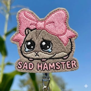 Puede incluir: Una insignia bordada gris con un hámster triste con ojos grandes y llorosos y un lazo rosa. El texto "SAD HAMSTER" está bordado debajo. La insignia tiene un clip retráctil.