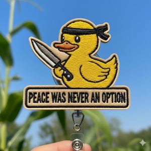 May include: Embroidered badge of a yellow duck with a black headband, holding a knife. The badge has the text "PEACE WAS NEVER AN OPTION" in a rectangular frame. The badge is attached to a retractable reel.