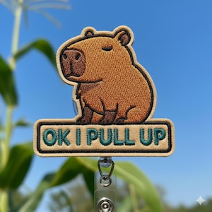 May include: An embroidered badge featuring a brown capybara with the text "OK I PULL UP". The text is in green letters. The badge has a light tan border and a silver retractable badge reel.