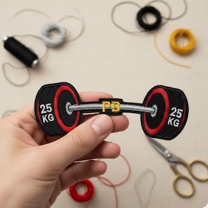 Peut inclure: Écusson brodé représentant une barre de musculation avec des poids noirs et des détails rouges. Les poids indiquent "25 KG". La barre porte les lettres "PB" en or. L'écusson est tenu dans une main.