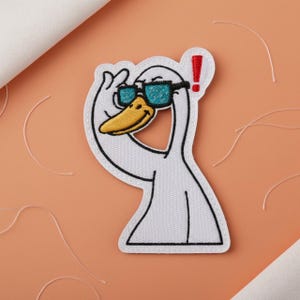 May include: Embroidered patch of a white cartoon goose wearing black sunglasses with teal lenses. The goose is giving a thumbs up and has a yellow beak. A red exclamation mark is next to the goose's head. The patch has a white border.