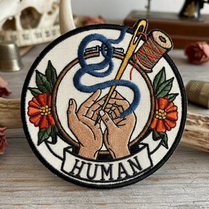 May include: A circular patch with a cream background features a needle and thread design. The needle is gold, and the thread is blue. A spool of colourful thread sits next to the needle. The word "HUMAN" is printed on a banner below the design. Two orange flowers with green leaves are on either side.