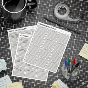 Pode incluir: Uma vista aérea de um espaço de trabalho organizado com blocos de notas "ADHD BRAIN DUMP", canetas, uma caneca de café e fita adesiva. Os blocos de notas têm seções para tarefas, compromissos e notas. Um frasco de vidro contém canetas de várias cores.