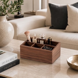 Puede incluir: Un organizador rectangular de madera marrón oscuro con cinco compartimentos interiores de distintos tamaños. El organizador contiene una brocha de maquillaje, un frasco cuentagotas, un frasco de perfume cuadrado, un labial negro y un pequeño estuche compacto.