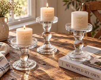 May include: Three clear glass candle holders with tiered, rounded stems and wide circular bases. Each holder supports a white pillar candle labeled "SALT CANDLE classic" with a lit wick, creating a warm glow on the wooden surface.