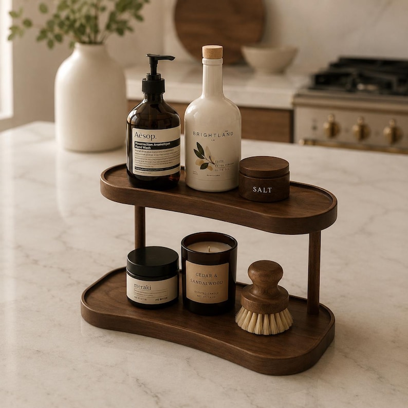 May include: A two-tiered oval wooden organizer shelf holding a dark amber Aesop hand wash bottle, a white Brightland olive oil bottle, a small dark brown salt jar, a Meraki jar, a Cedar & Sandalwood candle, and a round wooden dish brush.
