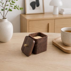 May include: A square dark brown wooden box with a matching lid featuring a small gold-toned knob. The box is open, revealing a collection of light-coloured wooden toothpicks inside. The container has rounded corners and a smooth, polished wood grain finish.