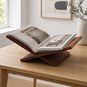 Könnte beinhalten: Ein dunkelbrauner hölzerner Buchständer mit X-förmigem Fuß, der ein aufgeschlagenes Hardcover-Buch mit schwarz-weißen Innenseiten hält. Der Ständer verfügt über zwei angewinkelte Platten, die das Buch in einer offenen Position stützen.