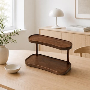 May include: A two-tiered wooden display stand featuring a dark brown finish with smooth, rounded edges. The stand consists of two oval-shaped trays connected by two vertical supports, creating an open structure for organizing or displaying items.