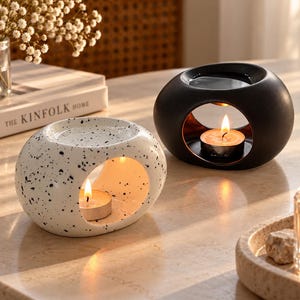 Può includere: Due bruciatori di essenze in ceramica di forma ovale, uno nero opaco e uno bianco con macchie nere. Ogni bruciatore presenta un'apertura centrale per una candelina e un piattino poco profondo sulla parte superiore.