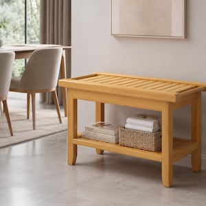 Puede incluir: Un banco de madera de color marrón claro con una superficie superior de listones y un estante inferior. El estante inferior contiene una pequeña cesta tejida con toallas blancas dobladas y una pila de dos libros con texto visible.