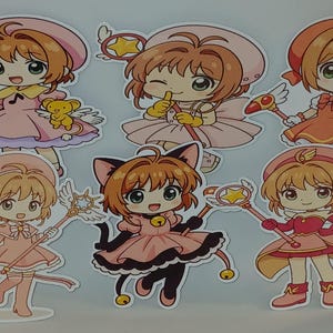 Puede incluir: Una colección de seis pegatinas troqueladas con personajes de estilo anime. Cada personaje tiene ojos grandes y viste un atuendo diferente en tonos de rosa, rojo y negro. Algunos personajes sostienen varitas o tienen alas.
